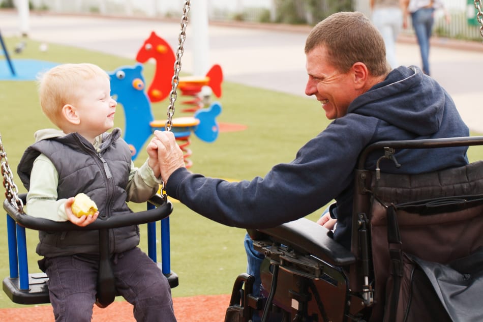 Picture of a dad in a wheelchair holding hands with his son who is on the swing. 