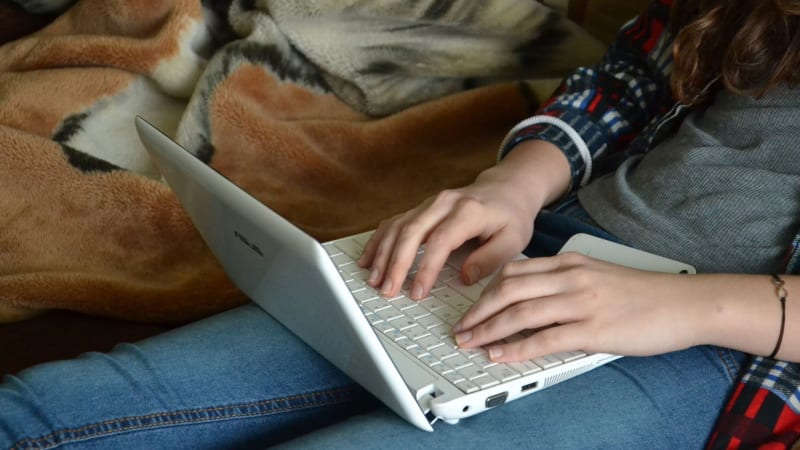 A girl sits on a bed typing on a white laptop. 