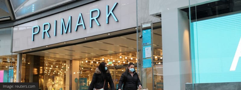 A photograph of a Primark shop. A grey building with a blue Primark sign and two shoppers entering the store with bags in their hand. The image is sourced from reuters.com.