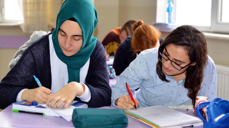 A young student wearing a blue hijab sits at a desk next to her friend. They are both writing. 