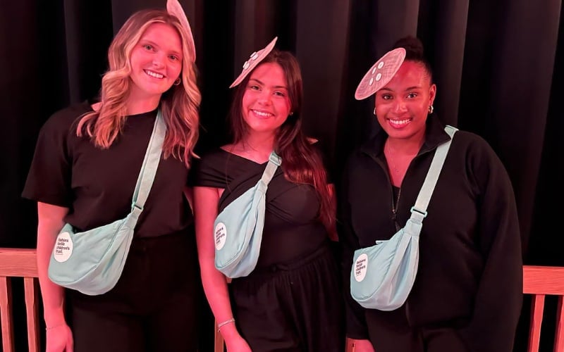 Three female volunteers stand against a black background. They are in their early twenties and are wearing blue bags and pink fascinators. 
