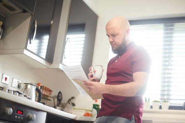 A white male with a beard and shaved head, stands in his kitchen. The kitchen has grey cabinets and a large window. The man is holding a household bill in his hand and is looking worried. 