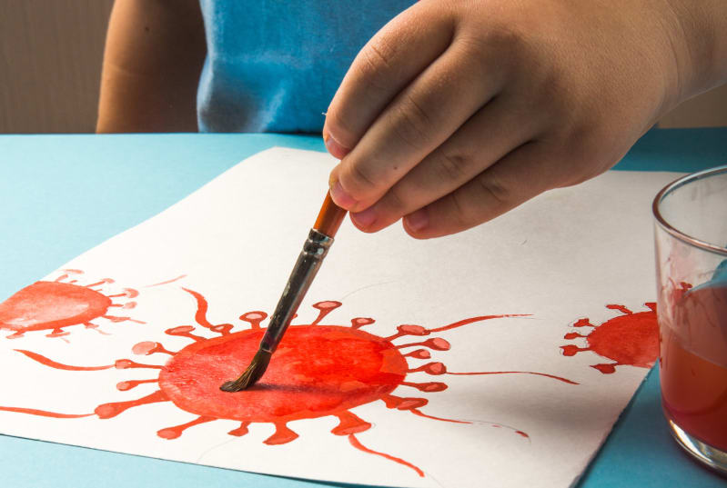 A child in a blue tshirt paints a coronavirus cell in red paint.